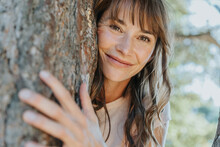 Smiling Mature Woman Leaning On Pine Tree In Public Park