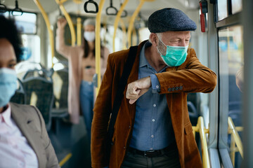 Wall Mural - Mature man with face mask sneezing into elbow while commuting by bus.