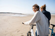 © UWE_UMSTAETTER/Westend61 - Smiling beautiful woman walking with bicycle by boyfriend while looking over shoulder at beach against clear sky on sunny day
