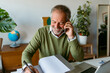 © VALENTINA BARRETO STUDIO/Westend61 - Smiling man writing in book while talking on mobile phone at home