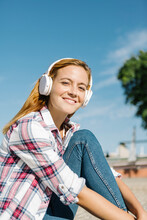Smiling Woman Listening Music On Headphone Sitting On Footpath During Sunny Day
