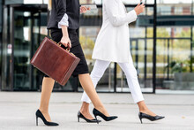 Young Businesswoman With Briefcase Using Mobile Phone While Walking By Colleague On Footpath
