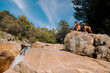 © Manu Reyes, www.vientocuatro.com/Westend61 - Trekker sitting with dog on rock by waterfall in forest at La Pedriza, Madrid, Spain