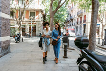 Female Friends Talking While Walking On Sidewalk In City