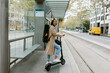 © VALENTINA BARRETO STUDIO/Westend61 - Woman with push scooter and instrument case standing at tramway station in city
