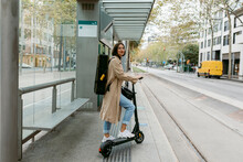 Woman With Push Scooter And Instrument Case Standing At Tramway Station In City