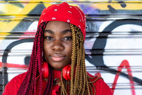 Plakat Portrait of an exotic black girl with colored braids. Red headphones hanging from the neck. Dressed in a red sweater and a red headscarf. Graffiti wall background