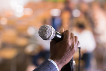 Closeup of microphone in hand of African American man on blurred background of conference room. Concept of speaking during public event