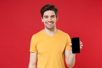 Young smiling unshaved caucasian cheerful man in casual yellow t-shirt hold mobile cell phone centre with blank screen workspace copy space mock up isolated on red color background studio portrait