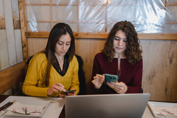 Close-up two young attractive women sitting at table restaurant while using computer laptop and holding smartphone. Lunch break, freelance work concept.