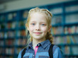© Comeback Images - Head and shoulders portrait of beautiful blonde little girl with braided hair and freckles smiling at camera standing near book shelves in school library