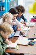 © Comeback Images - Side view of elementary school girl doing task on laptop computer helped by female teacher, her defocused classmates studying sitting at desk in school classroom nearby