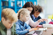 © Comeback Images - Side view selective focus of elementary school girl doing task on laptop computer helped by female teacher, her defocused fellow students studying sitting at desk in school classroom nearby