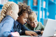 © Comeback Images - Group of three excited diverse students using laptop sitting at desk in school library. Mixed race schoolboy and his friends browsing through information on Internet
