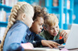 © Comeback Images - Group of three curious diverse kids using laptop sitting at desk in school library. Mixed race schoolboy and his friends browsing through information on Internet
