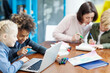 © Comeback Images - Two elementary school kids, boy and girl, having fun browsing through pictures on laptop computer sitting at desk in classroom, while teacher helping their classmate in background