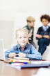 © Comeback Images - Beautiful blonde little girl looking at camera while writing in notepad sitting at desk in school classroom with open laptop in front of her, two defocused classmates talking in background