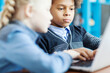 © Comeback Images - Side view of two elementary students, boy and girl, working on laptop computer sitting at desk in school library. Focus on serious curly-haired mixed race boy typing on keyboard