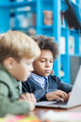 Side view of two boys working on laptop computer sitting at desk in school library. Focus on curly-haired mixed race boy typing on keyboard