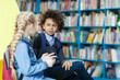 © Comeback Images - Three students - blonde boy, mixed race curly-haired boy and blonde girl with braids - sitting on desk in school library and talking, schoolgirl telling story to male friends