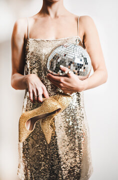 Young woman figure in festive glittering cocktail golden mini dress holding discoball and gold high heels shoes in hands over white wall. New Year holiday party concept