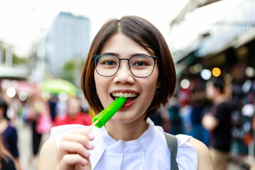 Asian young short hair beautiful woman wearing glasses enjoy eating green popsicle or ice cream at street market, street food Thailand