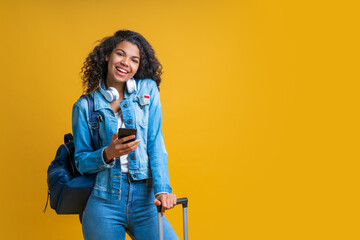 Wall Mural - Portrait of cute smiling dark skinned girl posing with mobile phone,  hand luggage bag and backpack, isolated over bright yellow background