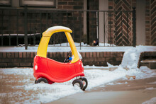 Red And Yellow Toy Plastic Ride In Car Parted By Sidewalk In Front Of House In Snow - Safe Driving - Selective Focus