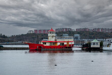 Urban Winter Scene With A Red Lighthouse Light Ship Houseboat With Dramatic Clouds And Buildings In The Background.