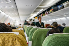 Commercial Plane Interior Before Take Off, Focus On Man Head At Seat Ahead, Most Of Passengers Already Seated, Baggage Compartments Still Opened, Blurred Stewardess In Background
