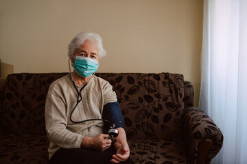 Portrait of a caucasian senior woman with a face mask measuring her blood pressure with a pressure gauge in her house. Health check during the COVID-19 coronavirus pandemic