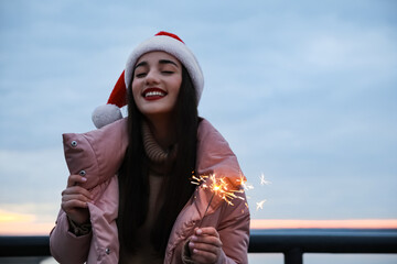 Wall Mural - Woman in Santa hat and warm clothes holding burning sparkler outdoors