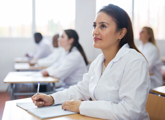positive latin american girl in white coat listening to lecture and taking notes in classroom during