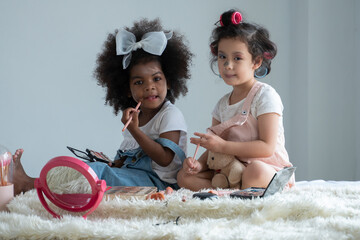 two little girls, african and asian children enjoy playing mother’s cosmetic make up together on bed