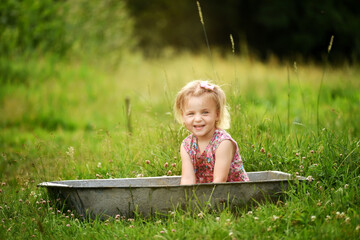 A little girl in a dress splashes in a trough of water in the middle of a green meadow. Rural cute photo of a carefree childhood.