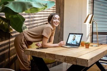 Wall Mural - Portrait of a young and cheerful woman in domestic suit having a video connection with colleagues, working on laptop from a cozy home office