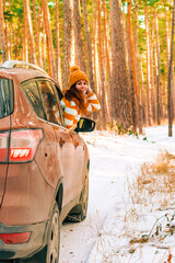 A smiling woman in a sweater and hat looks out of the car window against the background of a snowy landscape in a winter pine forest, road trip