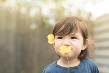A Toddler With A Yellow Flower On Her Hair Holds Another Yellow Flower In Front Of Her Face And Smiles As She Smells It In The Garden Of A House In Edinburgh, Scotland, United Kingdom