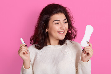 Photo of attractive Caucasian woman holds cotton tampon and sanitary napkin in hands, has happy look, monthly menstrual cycle, wears white sweater. Protective care for women health.