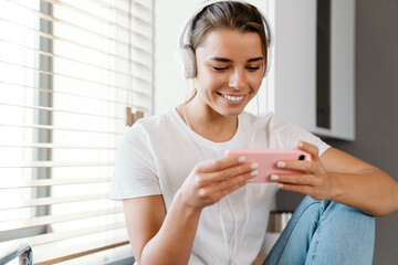 Wall Mural - Beautiful cheerful girl in headphones using mobile phone while sitting