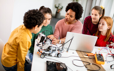 Happy kids with their African American female science teacher with laptop programming electric toys and robots at robotics classroom