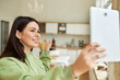 © Anatoliy Karlyuk - Portrait of dark haired positive young female chatting with friend using online app for video chat on touchscreen pad, pointing finger at kitchen in background, showing new furniture, smiling
