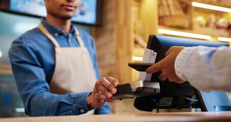 Close up of African American male seller in face mask and protective black gloves selling baked fresh bread in bakery shop. Client paying with credit card on device buying baking. Business concept