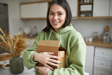 Wall Mural - Portrait of beautiful young dark haired female in stylish hoodie carrying carton box, unpacking her things after moving yo new apartment, looking at camera with joyful smile, posing in kitchen