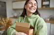 Adorable emotional young female in comfy clothes having excited facial expression, smiling happily, has just got home delivered parcel, enjoying online shopping, purchasing new clothes on sale