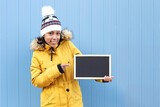 Fototapeta Panele - Portrait of a hispanic girl wearing winter clothes holding and pointing at a blank chalkboard. She is standing next to a blue wall. Space for text.