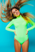 Attractive Young Slim Female Model With Flying Long Fair Hair Dressed In Bright Green Bodysuit Standing Against Blue Background