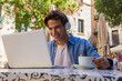 © David Munoz/ADDICTIVE STOCK - Overjoyed young male in wireless headphones getting incredible news on laptop and celebrating victory while sitting at table with cup of coffee on cafe terrace