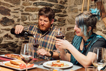 Content Friends Sitting At Wooden Table With Dishes And Pouring Refreshing Red Wine In Glasses While Having Dinner In Restaurant