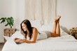 © Gustavo Pozo/ADDICTIVE STOCK - Cheerful millennial female in nightwear lying on bed and surfing internet on laptop while spending weekend morning in cozy bedroom with minimalist interior